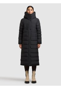 Must puffer-jope kõrge kraega, pikkuse ja tikitud tekstuuriga ning ees nööpidega. Kantud mustade retuuside ja beežide pahkluu saabastega.