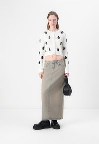 Donna che indossa un maglione corto bianco con motivi floreali neri, una lunga gonna di jeans beige, stivaletti neri chunky e tiene in mano una borsa nera.