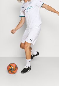 Bílý fotbalový dres s tyrkysovými akcenty, černé logo PUMA, v kombinaci s černými fotbalovými kopačkami a bílými ponožkami. V blízkosti se nachází oranžový a černý fotbal.