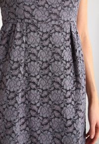 Robe en dentelle grise avec motif floral ; corsage ajusté et jupe évasée ; présente une texture délicate et des détails de design complexes.