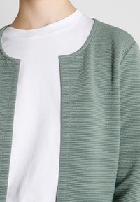 Ljusgrön, strukturerad jacka med rund halsringning, bärs över en vit skjorta. Har ett ribbat mönster med öppen framkant.