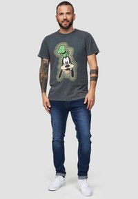T-shirt in cotone grigio scuro con una grafica colorata di Goofy. Indossato con jeans blu e sneakers bianche, con dettagli visibili di tatuaggi sulle braccia.