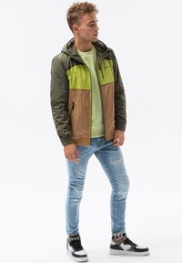 Färgblockerad, lätt jacka i olivgrön, grön och brun med huva; matchad med ljusblå slitna jeans och svartvita sneakers.