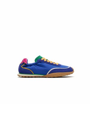 Zapatilla deportiva azul con cordones amarillos, lengüeta verde, lengüeta rosa en el talón y suela de goma, mostrada de perfil sobre fondo blanco.