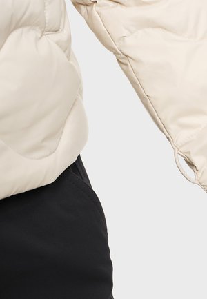 Manche d'une veste beige matelassée avec un poignet élastique et pantalon noir visible sur un fond clair.