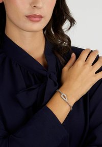 Bracciale in argento con un centro a forma di goccia, decorato con pietre trasparenti, montato su una catena a maglie, indossato su un polso.