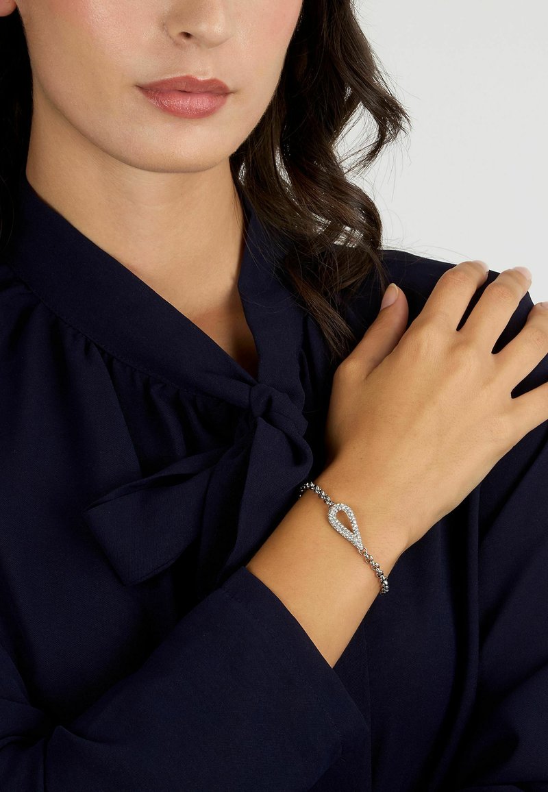 Bracciale in argento con un centro a forma di goccia, decorato con pietre trasparenti, montato su una catena a maglie, indossato su un polso.