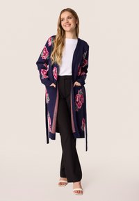 Marineblauwe lange cardigan met grote roze bloemenprints, open voorkant, strikceintuur en zakken, gecombineerd met zwarte broeken en witte sandalen.