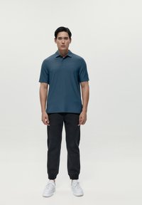 Polo da uomo a maniche corte in teal, realizzato in tessuto morbido, abbinato a joggers neri affusolati e sneakers bianche su uno sfondo neutro.