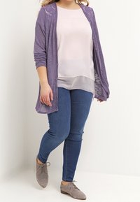 Lilla strikket cardigan over en lys pink bluse, parret med blå skinny jeans og grå snørebåndssandaler, med en afslappet pasform og knapdetaljer.