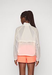 Veste blanche légère et transparente avec un col montant, design superposé et poignets élastiques. Associée à un short orange néon.