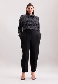 Pantaloni neri con pieghe sartoriali abbinati a una camicetta scura a pois con colletto a fiocco. Tessuto liscio con texture lucida.
