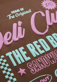 Barna textil, amelyen xilogravírozott szöveg található rózsaszín és világoskék színben. A szöveg tartalmazza a következőket: "Az Eredeti Deli Club otthona" és "$15", csillag- és négyzetmintákkal.