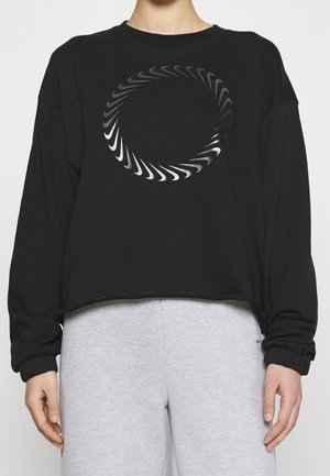 Personne portant un haut noir à manches longues avec un motif circulaire en spirale sur la poitrine et un pantalon gris clair.
