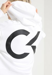 Sweat à capuche blanc avec un grand motif noir "C2" au dos, doté d'une texture douce et d'une capuche. Les manches sont ajustées avec des poignets côtelés.