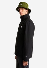 Mies pukeutuneena oliivinvihreään bucket-hattuun ja mustaan vetoketjutakkiin, jossa on korkea kaulus, seisoo sivuprofiilissa valkoista taustaa vasten.