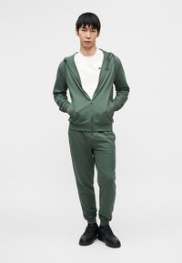 Roheline lukuga kapuutsiga dressipluus taskutega, sobitatud matching dressipükstega. Valge T-särk selle all. Mustad spordijalatsid. Pehme, sile kangatekstuur.