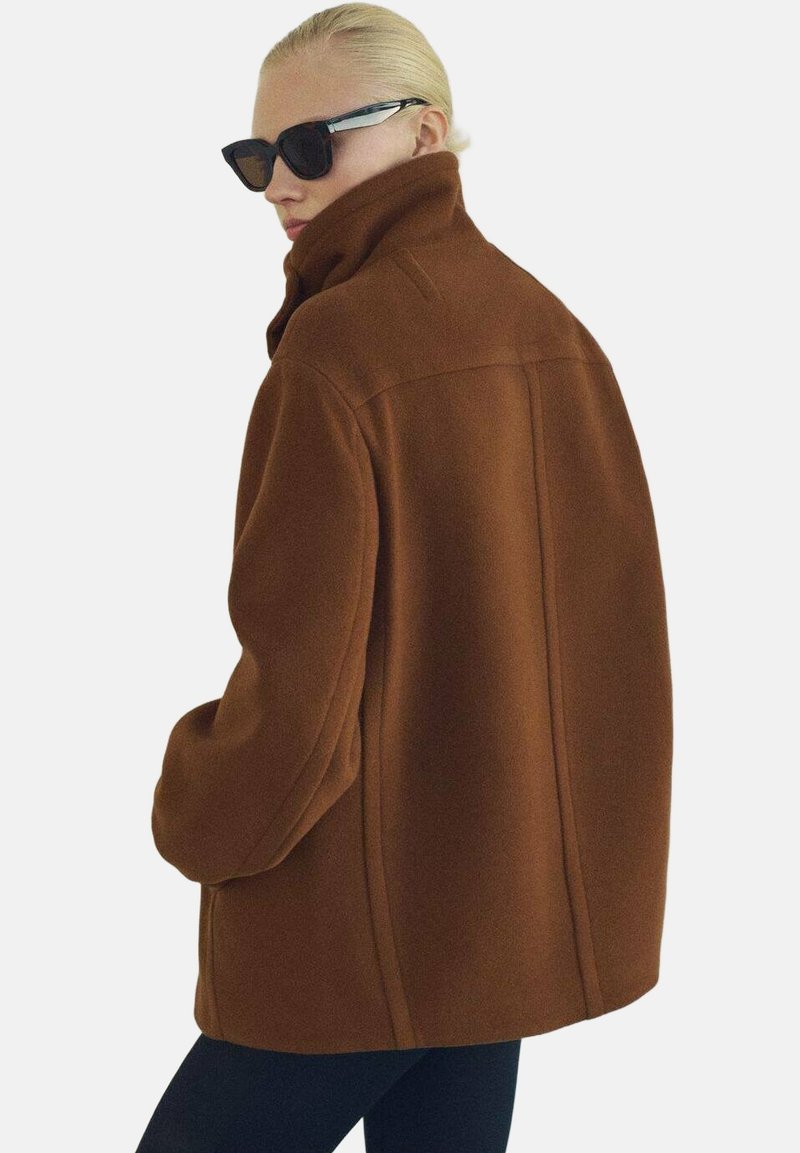 Hnědý kabát z vlněné směsi, oversized střihu s vysokým límcem, viditelnými švy a uvolněným střihem. Nosit s černými slunečními brýlemi a úzkými černými kalhotami.