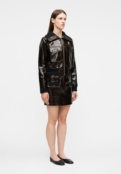 Γυναίκα όρθια με μια γυαλιστερή μαύρη ζακέτα με φερμουάρ και ταιριαστή φούστα με μπροστινές τσέπες, συνδυασμένη με μαύρα μπαλέτα.