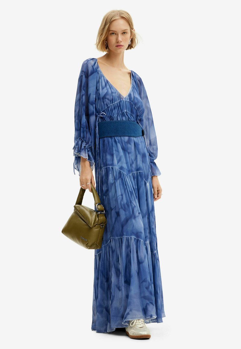 Maxikleid mit einem fließenden Design in verschiedenen Blautönen, langen Ärmeln und tailliertem Schnitt. Begleitet von einer olivgrünen Tasche.