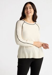 Hellbeige Pullover mit geripptem Saum, rundem Halsauschnitt und schwarzen Akzenten entlang der Schultern und Ärmel. Weiche Textur, lange Ärmel.