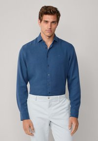 Uomo che indossa una camicia a maniche lunghe blu infilata nei pantaloni grigio chiaro, in piedi contro uno sfondo semplice.