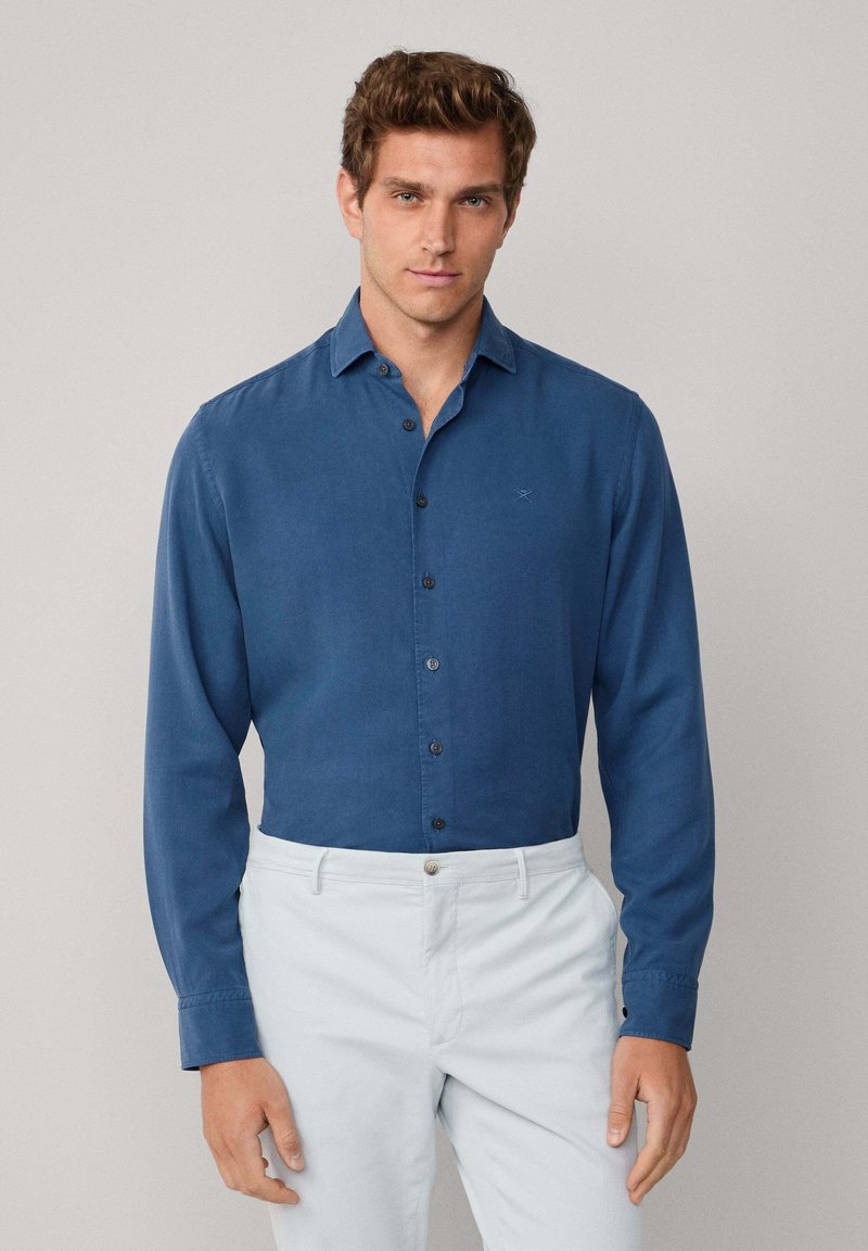 Uomo che indossa una camicia a maniche lunghe blu infilata nei pantaloni grigio chiaro, in piedi contro uno sfondo semplice.