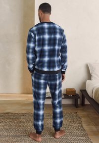 Bărbat desculț, stând pe un covor țesut, purtând un top cu mânecă lungă în carouri albastre și albe și pantaloni asortați, într-un dormitor minimal.