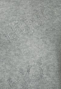 Maglione grigio chiaro a maglia con lettere in rilievo e decorazioni argentate, che creano un design testurizzato su tutto il tessuto.