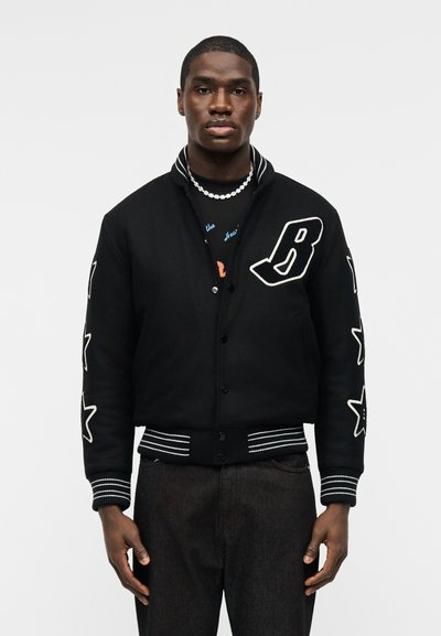 Άνδρας που φοράει μαύρο varsity μπουφάν με άσπρο γράμμα "B" στο στήθος και άσπρα αστέρι σχέδια στα μανίκια, συνοδευόμενο από σκούρο παντελόνι και κολιέ με χάντρες.