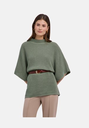 Femme aux cheveux bruns portant un pull tricoté vert à manches courtes cintré avec une ceinture marron, un pantalon beige et des boucles d'oreilles créoles dorées.