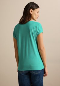 Top de manga corta turquesa con detalle de hombros fruncidos, fabricado en tejido suave, combinado con jeans de denim azul. Se muestra la vista trasera.