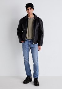 Chaqueta de cuero negra, camiseta verde oliva, vaqueros azules y botas negras con cordones. El modelo también lleva un gorro negro y un collar de cuentas.