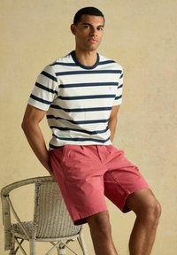 Bărbat tânăr stând pe marginea unui scaun de nuiele, purtând o cămașă cu mâneci scurte în dungi albă și bleumarin și pantaloni scurți corali, pe un fundal bej.