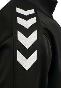 Schwarzer Jackenärmel mit vier weißen, nach unten zeigenden Chevronen mit dezenten sich wiederholenden „K“-Mustern.