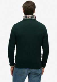 Suéter de punto verde oscuro con cuello y patrón de cuadros, usado sobre una camisa. Detalles de costura visibles y puños de canalé.
