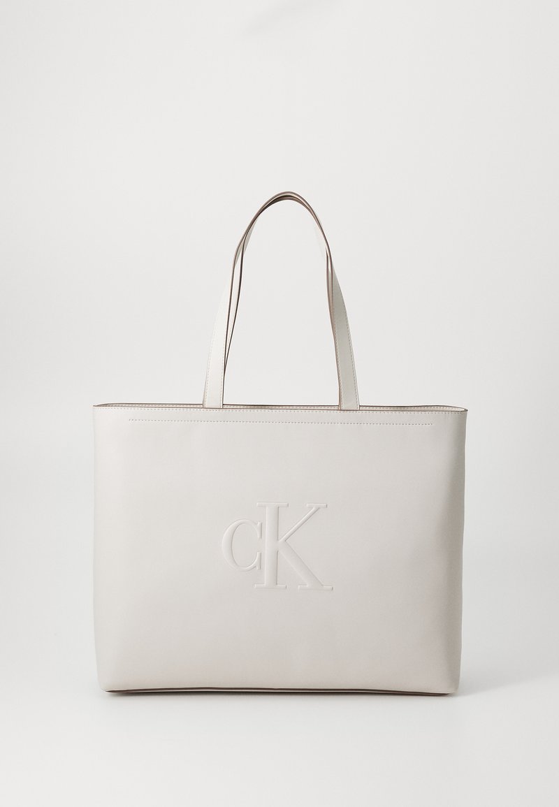 Grande borsa tote in pelle beige chiaro con logo CK in rilievo, manici piatti e design minimalista, caratterizzata da una superficie liscia e senza hardware visibile.