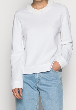 Osoba nosząca prosty biały sweter z długim rękawem z prążkowanym kołnierzem oraz jasnoniebieskie dżinsy z wysokim stanem, ręka w kieszeni.