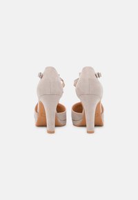 Beige suede high-heeled shoes with an open back, adjustable ankle strap, and a smooth texture. Wooden-like heels with a subtle curve.