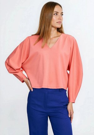 Koralowa bluzka z dekoltem w serek i luźnymi rękawami, wykonana z teksturowanej tkaniny, w połączeniu z dopasowanymi, niebieskimi spodniami. Gładkie wykończenie, krótka długość i subtelne przeszycia.