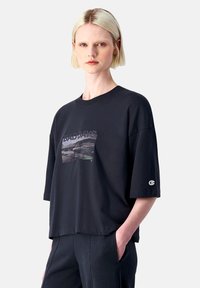 Schwarzes T-Shirt mit kurzen Ärmeln aus Baumwolle im Oversized-Fit. Mit einem grafischen Druck auf der Vorderseite, einem gerippten Halsausschnitt und einem Logo auf dem Ärmel.