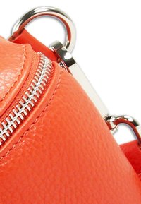 Oranje leren tasgedeelte met een metalen rits, gladde textuur en zilveren hardware. Beschikt over een ronde ring voor bevestiging.