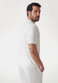 White polo shirt with short sleeves, textured side panels, minimalistic design, soft fabric, worn with matching white trousers.