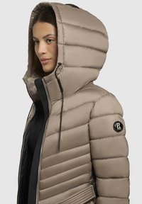 Beige pufferjack met een grote capuchon, verticale stiksels, ritsen en een badge op de linkermouw. Zachte textuur en slank ontwerp.