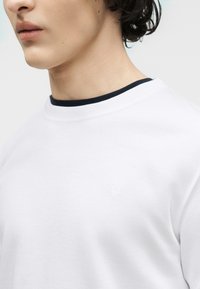 Νεαρό άτομο με σγουρά σκούρα μαλλιά, φορώντας ένα λευκό μπλουζάκι με στρογγυλή λαιμόκοψη και λεπτή μαύρη λεπτομέρεια στο γιακά, απλό φόντο.