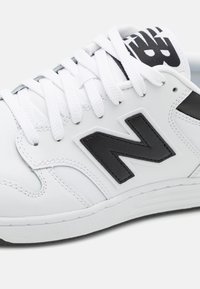 Baskets en cuir blanc avec des accents noirs, présentant un logo noir "N" prominent, des lacets blancs et une semelle en caoutchouc texturée.