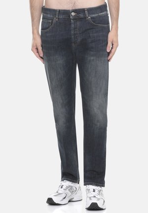 Uomo che indossa jeans blu scuro con sbiadimenti sulle cosce e scarpe da ginnastica bianche, in piedi contro uno sfondo neutro.