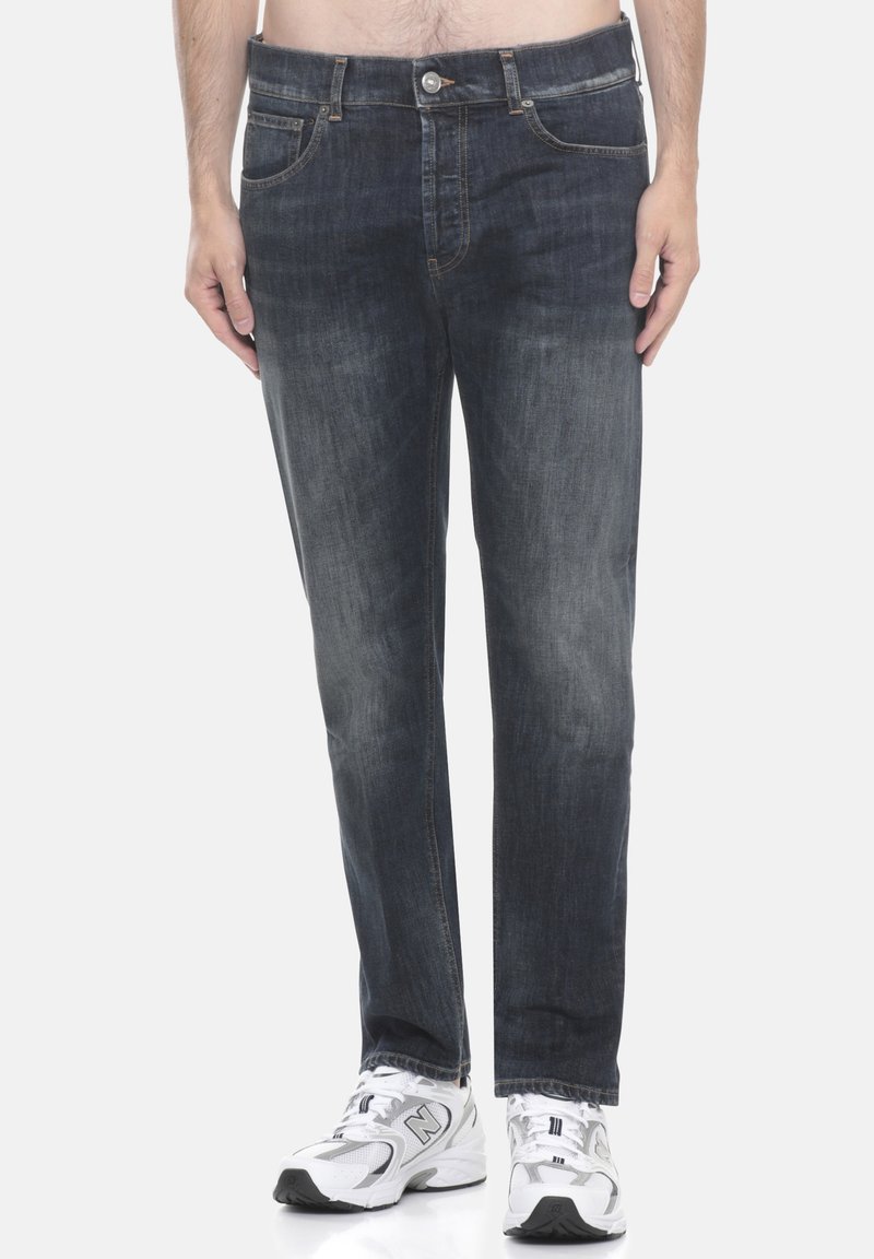 Uomo che indossa jeans blu scuro con sbiadimenti sulle cosce e scarpe da ginnastica bianche, in piedi contro uno sfondo neutro.