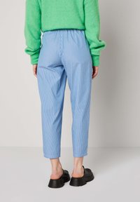 Henkilö, jolla on yllä sinivalkoiset raidalliset polvipituiset housut, kirkkaan vihreä neule ja mustat liukukengät paksulla pohjalla, seisoo koruttomalla taustalla.