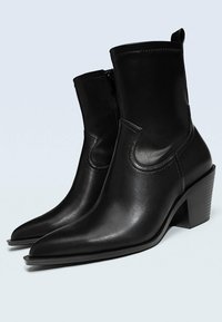 Botines de cuero negro hasta el tobillo con punta afilada, cierre de cremallera y tacón cuadrado. Textura suave con detalles de costura visibles.